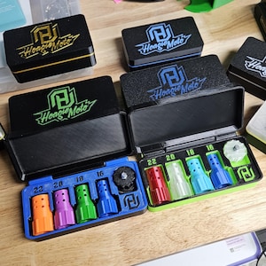 May include: Black rectangular cases with the "Hoagie Moto" logo, containing colorful nozzles labeled with sizes. The cases are open, revealing the contents. A green, 3D-printed holder and other tools are visible in the background.
