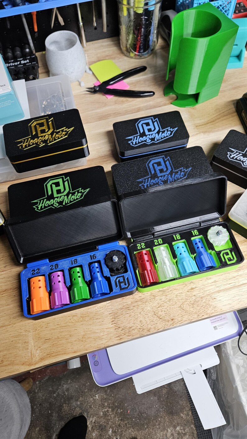 May include: Black rectangular cases with the "Hoagie Moto" logo, containing colorful nozzles labeled with sizes. The cases are open, revealing the contents. A green, 3D-printed holder and other tools are visible in the background.