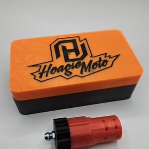 May include: An orange and black rectangular box with the text "Hoagie Moto" and a logo on the lid. A red and black cylindrical object with a metal fitting is next to the box. The objects are on a white surface.