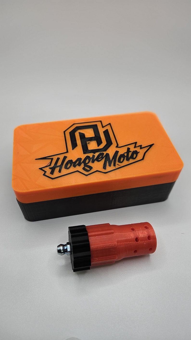 May include: An orange and black rectangular box with the text "Hoagie Moto" and a logo on the lid. A red and black cylindrical object with a metal fitting is next to the box. The objects are on a white surface.