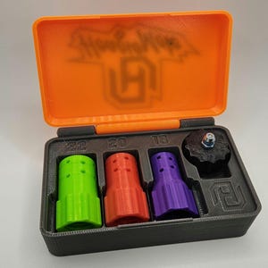 May include: An open, orange and black case containing several 3D-printed components. The case lid has the text "Show Cubes" and a logo. Inside are green, red, and purple cylindrical pieces, and a black knob with a bolt.
