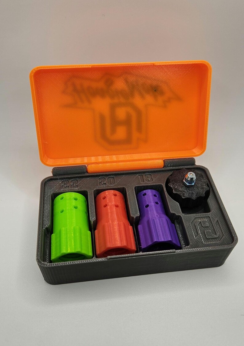 May include: An open, orange and black case containing several 3D-printed components. The case lid has the text "Show Cubes" and a logo. Inside are green, red, and purple cylindrical pieces, and a black knob with a bolt.