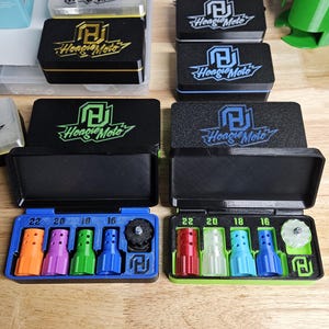 May include: Several black boxes with the "Hoagie Moto" logo, some with colored trim. Below, two blue trays hold colored nozzles labeled 22, 20, 18, and 16. The background includes tools and other boxes.