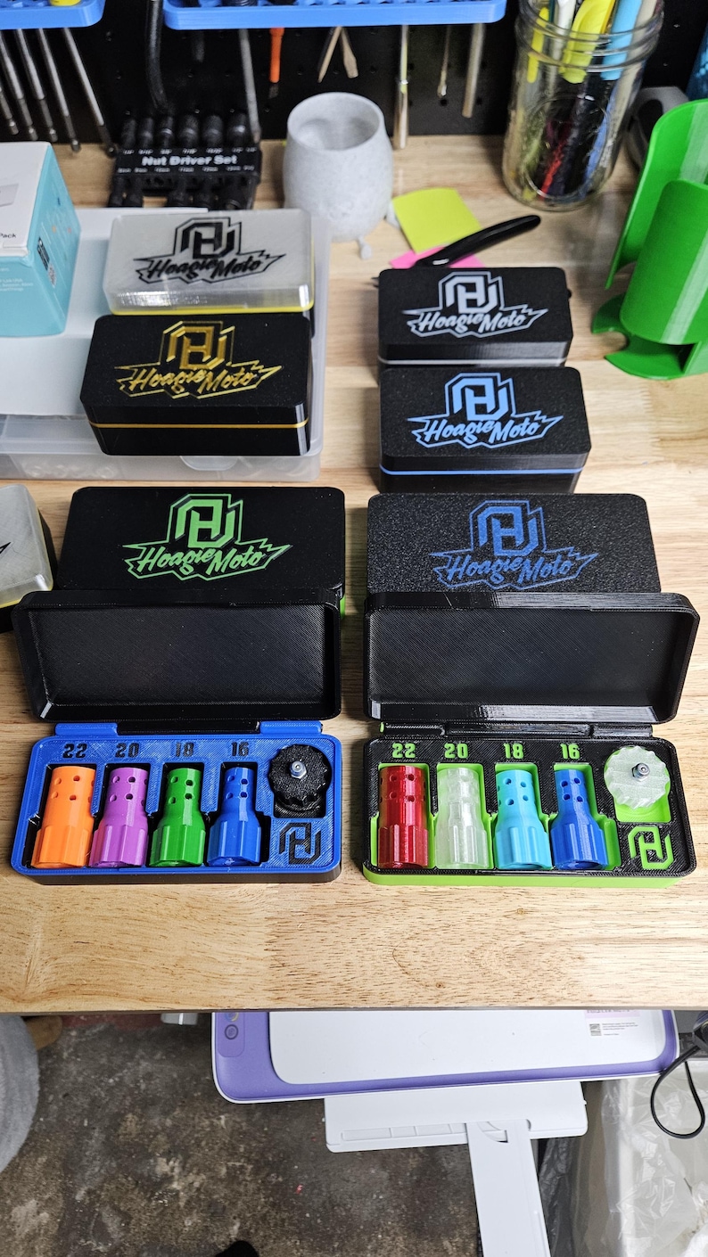 May include: Several black boxes with the "Hoagie Moto" logo, some with colored trim. Below, two blue trays hold colored nozzles labeled 22, 20, 18, and 16. The background includes tools and other boxes.