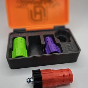 May include: A set of 3D-printed accessories in a black and orange case. The case contains a green, a black, and a purple piece. A red piece with a black end and a metal fitting is in front of the case.