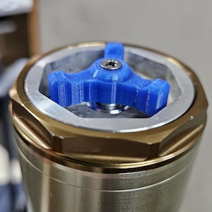 May include: Close-up of a blue, three-pronged tool inserted into a silver and gold metal component. The tool has a small screw in the center. The component has a hexagonal shape and is part of a larger mechanical assembly.