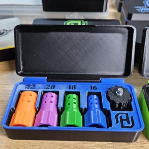 May include: A blue and black case with an open lid, containing four colorful, cylindrical objects in orange, purple, green, and blue. The case also holds a black, circular object. Numbers 16, 18, 20, and 22 are printed above the objects.