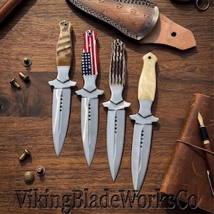 May include: Four fixed-blade knives with double-edged steel blades and distinct handles, including one with an American flag pattern, two with textured light-colored grips, and one with a smooth cream handle, alongside a leather sheath and pen.