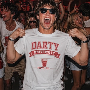 Può includere: Una maglietta bianca con la scritta "DARTY UNIVERSITY" in rosso, una grafica di bicchiere rosso e "PHYS. ED." sotto. La maglietta è mostrata da una persona con occhiali da sole, circondata da una folla.