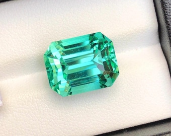 May include: A vibrant, emerald-cut gemstone with a bright, teal-green hue. The gem is set in a black jewelry box with a white interior. The facets of the stone reflect light, creating a sparkling effect.