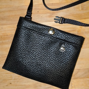 May include: A black faux leather pouch with a silver zipper pull and a silver charm. The pouch has a black adjustable strap with a plastic buckle.