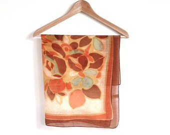 Peut inclure: Une écharpe à motifs floraux marrons, orange et crème suspendue à un cintre en bois sur un fond blanc.