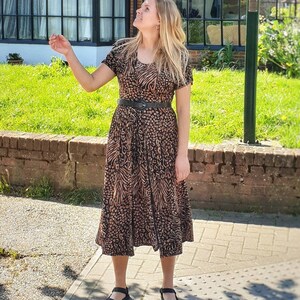 May include: A woman wearing a brown and black animal print midi dress with a black belt. She is standing on a sidewalk in front of a brick building with a large window.