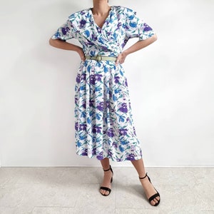 May include: A white dress with a purple and blue floral print. The dress has a V-neckline, short sleeves, and a fitted bodice. It has a gathered skirt and a belt.