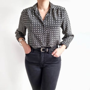 May include: A black and white patterned long-sleeved shirt with a collared neckline and button fastening. The shirt is tucked into black jeans with a silver belt.