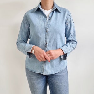 May include: A light blue denim shirt with embroidered details on the sleeves. The shirt has a button-down collar and long sleeves.