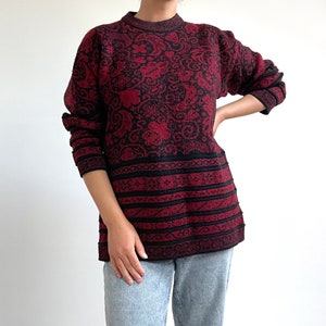 Op de afbeelding: Een rode en zwarte geruite trui met een ronde hals en lange mouwen. De trui heeft een bloemenpatroon en een gestreept patroon aan de onderkant.