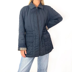 Op de afbeelding: Een donkerblauw gewatteerd jack met een ritssluiting en een geruite voering. Het jack heeft twee grote zakken aan de voorkant en een trekkoord in de taille.