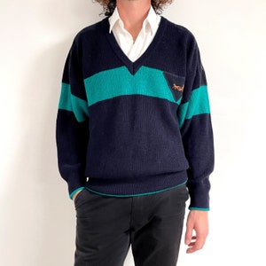 Könnte beinhalten: Ein marineblauer und türkisfarbener gestreifter Pullover mit V-Ausschnitt und einem kleinen gestickten Logo auf der Brust.