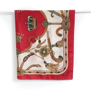 May include: A red silk scarf with a white background featuring a repeating pattern of gold and green crowns, swords, and tassels. The scarf has a gold border and the text "Fendi Roma" in the bottom right corner.