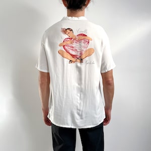 May include: White short-sleeved button-up shirt with a graphic print of a woman holding a heart. The text "Cavells Playa de Aro" is printed on the shirt.