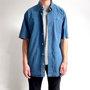 K&ouml;nnte beinhalten: Ein blaues, kurz&auml;rmeliges Hemd aus Denim mit einer Brusttasche.
