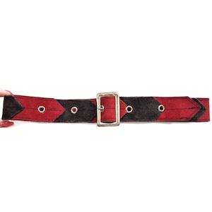 Può includere: Una cintura rossa e nera con fibbia color argento. La cintura presenta un motivo geometrico con sezioni rosse e nere. Ha occhielli metallici per la regolazione. La cintura è realizzata in materiale scamosciato.