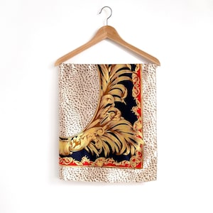 May include: A silk scarf with a cream leopard print background. The scarf has a detailed design with gold, navy blue, and red accents. It is hanging on a wooden hanger against a white background.