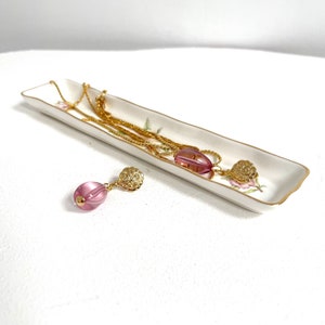 May include: A white ceramic dish with a floral pattern, holding a gold chain necklace with a pink glass pendant. A matching pair of earrings with pink glass beads and gold accents are on the white surface.