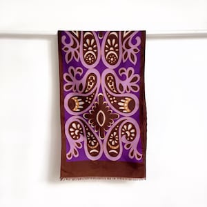 May include: A rectangular scarf featuring a repeating paisley pattern in shades of dark brown, light purple, and cream. The design includes symmetrical floral-inspired motifs and a solid dark brown border at the bottom with a fringed edge finish.