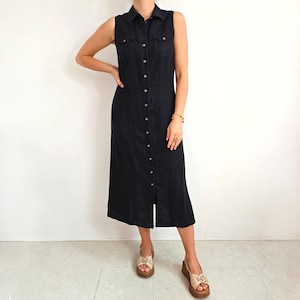 May include: A sleeveless black denim dress featuring a button-down front, two chest pockets, and a collared neckline. The dress extends to mid-calf length and has a small front slit. The model is wearing beige sandals.