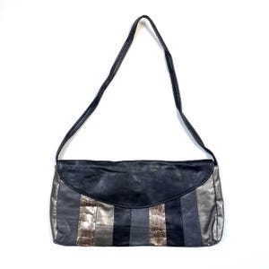 Op de afbeelding: Zwarte en zilveren schoudertas met een lange riem. De tas heeft een klepsluiting en een patchwork-ontwerp met afwisselende panelen van zwart, zilver en getextureerd bruin materiaal. De tas is rechthoekig van vorm.