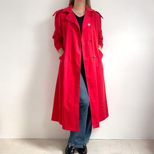 May include: A bright red trench coat with a double-breasted design and gold buttons. The coat has a classic collar, shoulder straps, and pockets. The long coat reaches the ankles and is worn with blue jeans and black shoes.