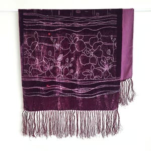 May include: Plum-colored velvet scarf with white floral embroidery and fringe. The scarf features a rectangular design with a solid purple border on one side. The bottom edge has a long fringe.