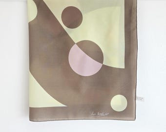 May include: A square scarf with a geometric design. The scarf features a pattern of brown, pale yellow, and pink shapes, including circles and diagonal lines. The scarf is made of a lightweight fabric and has a dark border. The designer's signature is visible.