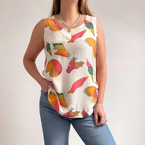 Puede incluir: Top sin mangas color crema con un estampado de conchas marinas en tonos rosa, naranja, amarillo y verde. El top tiene cierre de botones y cuello redondo. Se combina con vaqueros azules.