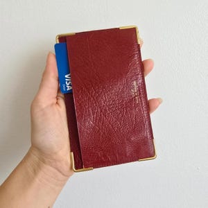 May include: A burgundy leather wallet with gold-colored corner accents. A blue Visa debit card is partially inserted into the wallet. The wallet has a textured surface and the text "CALF MADE IN ENGLAND".