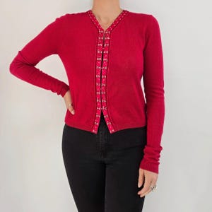 Puede incluir: Un cárdigan rojo vibrante con mangas largas. Los bordes delanteros están adornados con una cinta decorativa con un patrón repetido de pequeños adornos dorados. El cárdigan se combina con pantalones negros.