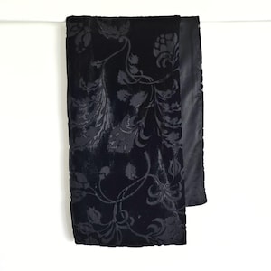 Op de afbeelding: Een zwarte sjaal met een bloemenpatroon. De sjaal is gemaakt van een zacht, fluwelig materiaal en heeft een herhalend ontwerp van bloemen en wijnranken. De randen zijn afgewerkt met een gladde, satijnachtige stof.