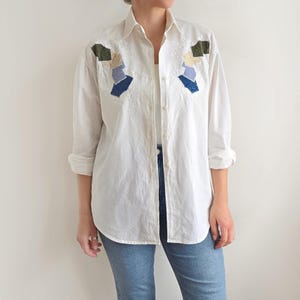 May include: White button-down shirt with embroidered floral details and patchwork accents in shades of blue, green, and beige. The shirt is paired with light blue jeans. The sleeves are rolled up.