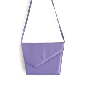 May include: A lavender-colored shoulder bag with a long, thin strap. The bag has a geometric design with a flap closure. The bag is made of a smooth material and is set against a white background.