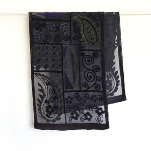 May include: A dark blue scarf with a patchwork design. The scarf features paisley, floral, and geometric patterns in shades of blue, gray, and black. The edges are trimmed with a dark border.