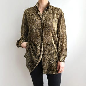 May include: A gold and black patterned button-down shirt. The long-sleeved shirt has a collared neckline and is partially unbuttoned. The shirt is worn with black trousers.