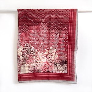 May include: A rectangular scarf with a floral garden design in shades of red, pink, and white. The scarf has a dark red border and a light gray trim. The design includes a watering can and various flowers and plants.