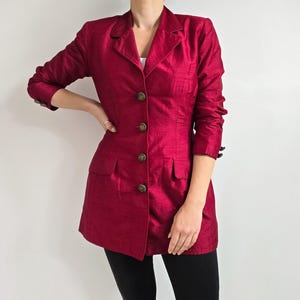 Puede incluir: Un blazer largo burdeos con un acabado texturizado. El blazer presenta un cuello con solapas, tres botones decorativos y dos bolsillos delanteros. Las mangas tienen detalles de botones en los puños. El blazer se usa sobre una camiseta blanca y se combina con pantalones negros.