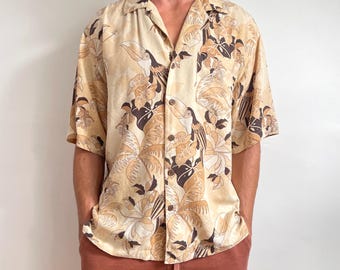 May include: A short-sleeved button-down shirt in a light beige colour featuring a repeating tropical print of toucans, palm leaves, and hibiscus flowers in shades of dark brown and cream. The shirt has a classic collar and a front button closure.