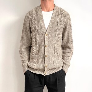 May include: A beige cable knit cardigan with a v-neck and button closure. The cardigan has two pockets on the front and a ribbed hem and cuffs.
