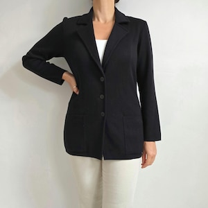 May include: A black blazer with a notched collar, two front pockets, and three buttons. The blazer is worn over a white top and paired with cream-coloured trousers. The blazer has long sleeves and a tailored fit.