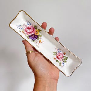 May include: A white rectangular ceramic dish with a gold trim and floral design. The floral pattern includes pink, purple, yellow, and blue flowers. The dish is held in a hand.