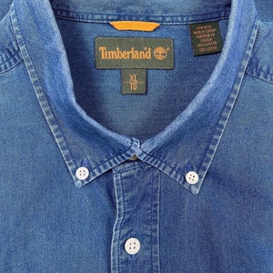 K&ouml;nnte beinhalten: Ein blaues Jeanshemd mit Knopfleiste und einem Timberland-Etikett. Das Etikett tr&auml;gt den Markennamen und die Gr&ouml;&szlig;e XL TG. Das Hemd ist in der T&uuml;rkei hergestellt und besteht aus 100% Baumwolle.
